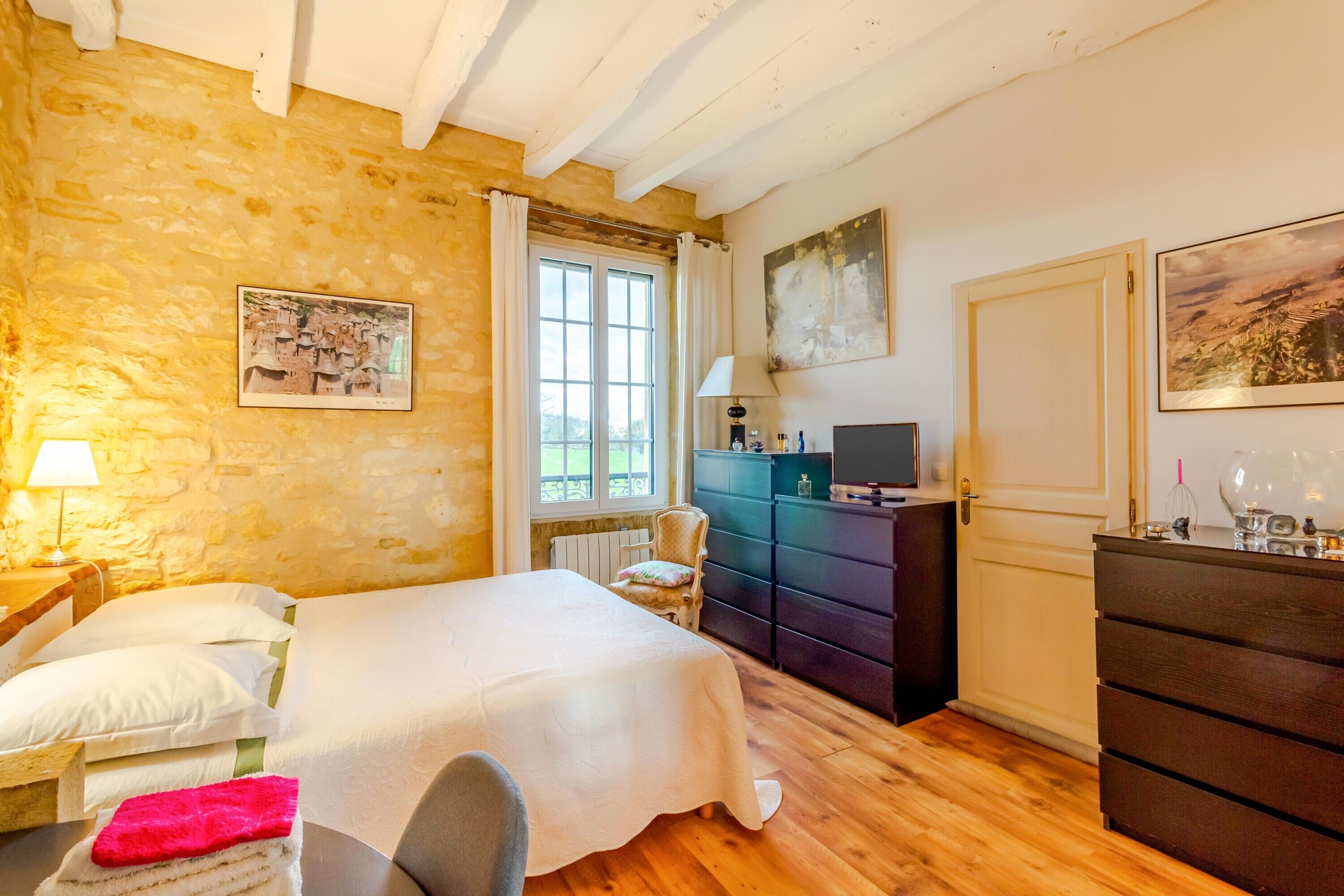 Een slaapkamer met een bed, een stoel, een dressoir, een televisie en een raam met gordijnen.