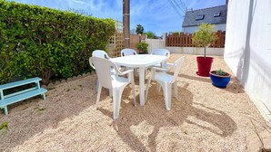 Un patio con una mesa y sillas blancas, rodeado de grava, setos y una cerca de madera.