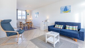 Una sala de estar con un sofá azul, una silla azul, una mesita auxiliar blanca y un comedor con mesa y sillas.