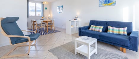 Ein Wohnzimmer mit einem blauen Sofa, einem blauen Sessel, einem weißen Beistelltisch und einem Essbereich mit Tisch und Stühlen.