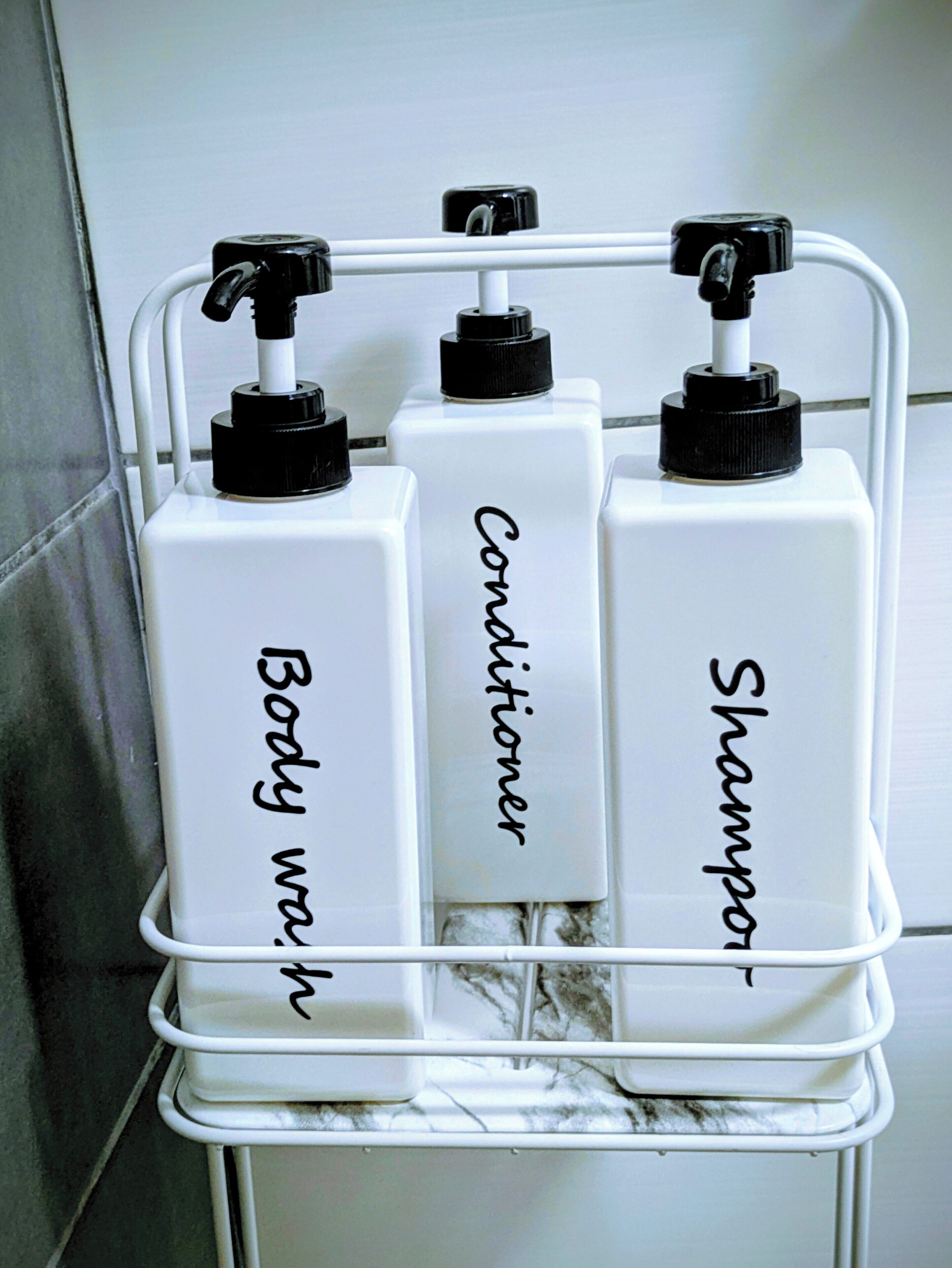 A bathroom shelf with three white bottles labeled "Body wash," "Conditioner," and "Shampoo."