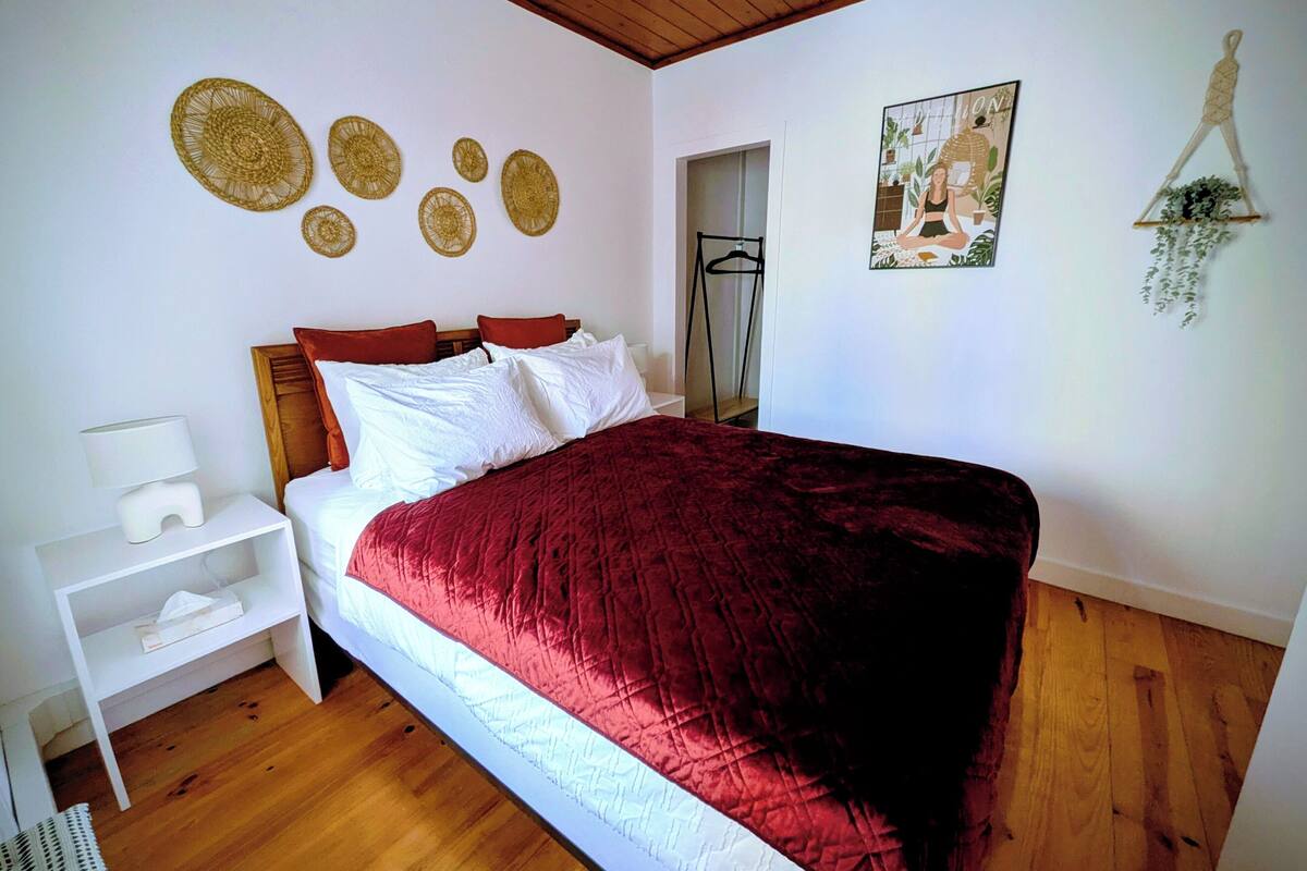 Un dormitorio con piso de madera, una cama con una manta roja, almohadas blancas, una mesita de noche con una lámpara y adornos en la pared que incluyen patrones circulares y un cuadro enmarcado.