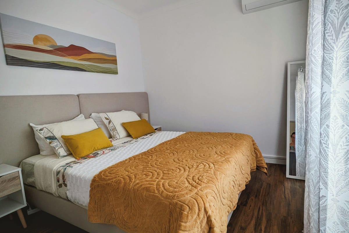 Un dormitorio con una cama, una mesita de noche, una lámpara, un cuadro en la pared y una ventana con cortinas.