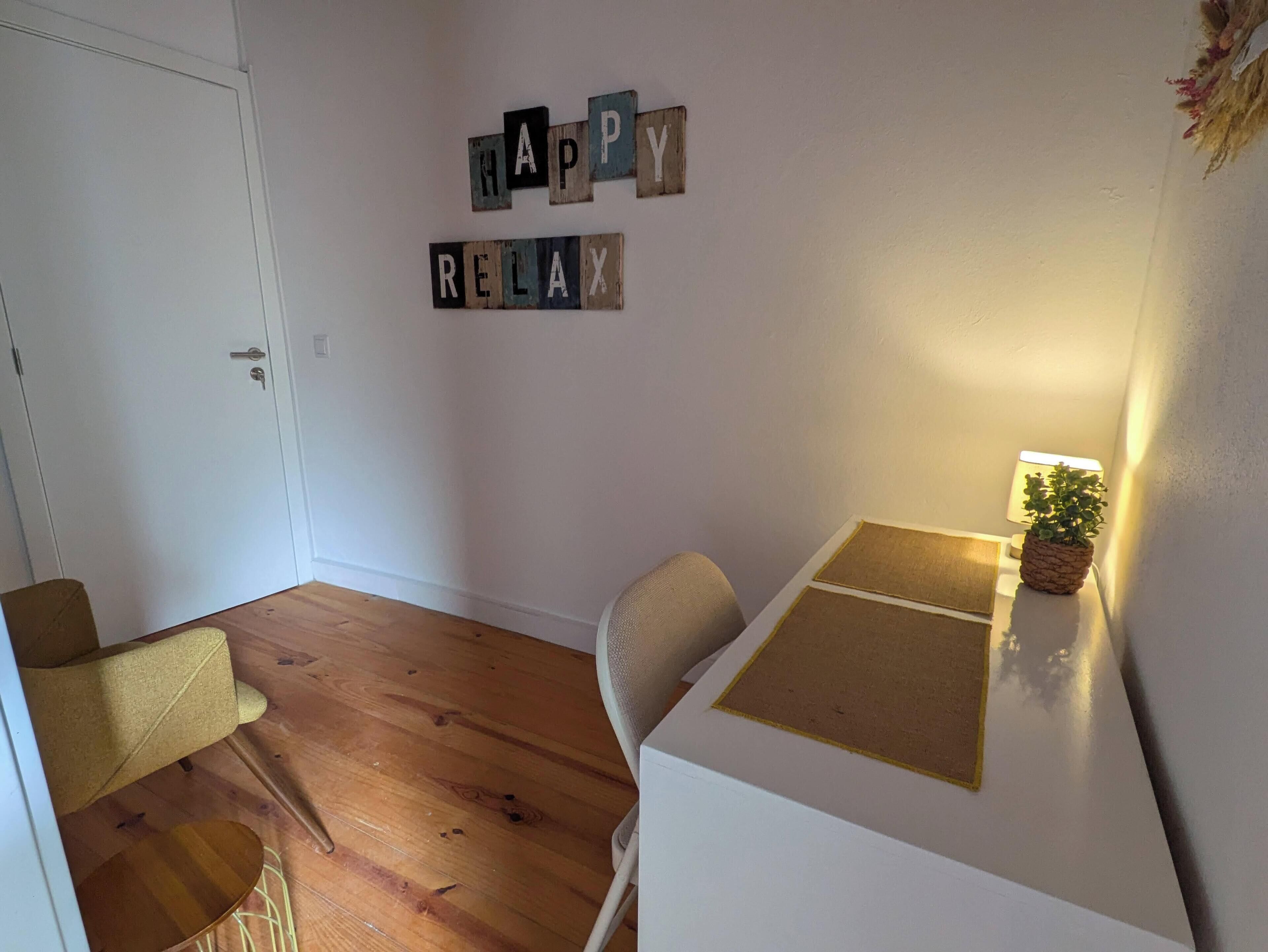 Une pièce avec un plancher en bois, une porte blanche, une petite table avec une plante et un mur sur lequel on a écrit « JOYEUSES VACANCES » à l’aide de blocs de bois.