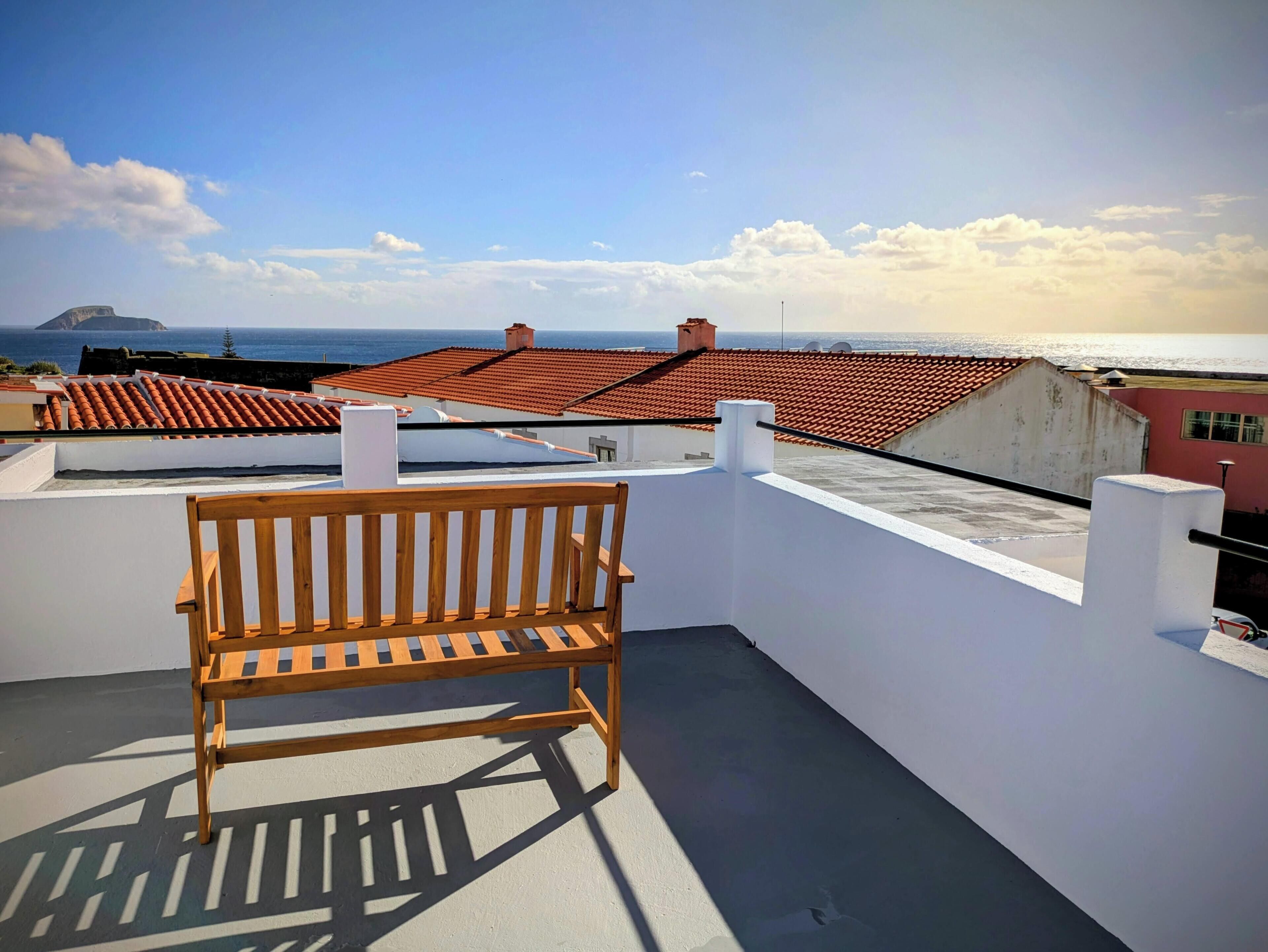 Un banco de madera en una terraza en la azotea, con vista al mar y tejados de tejas rojas.