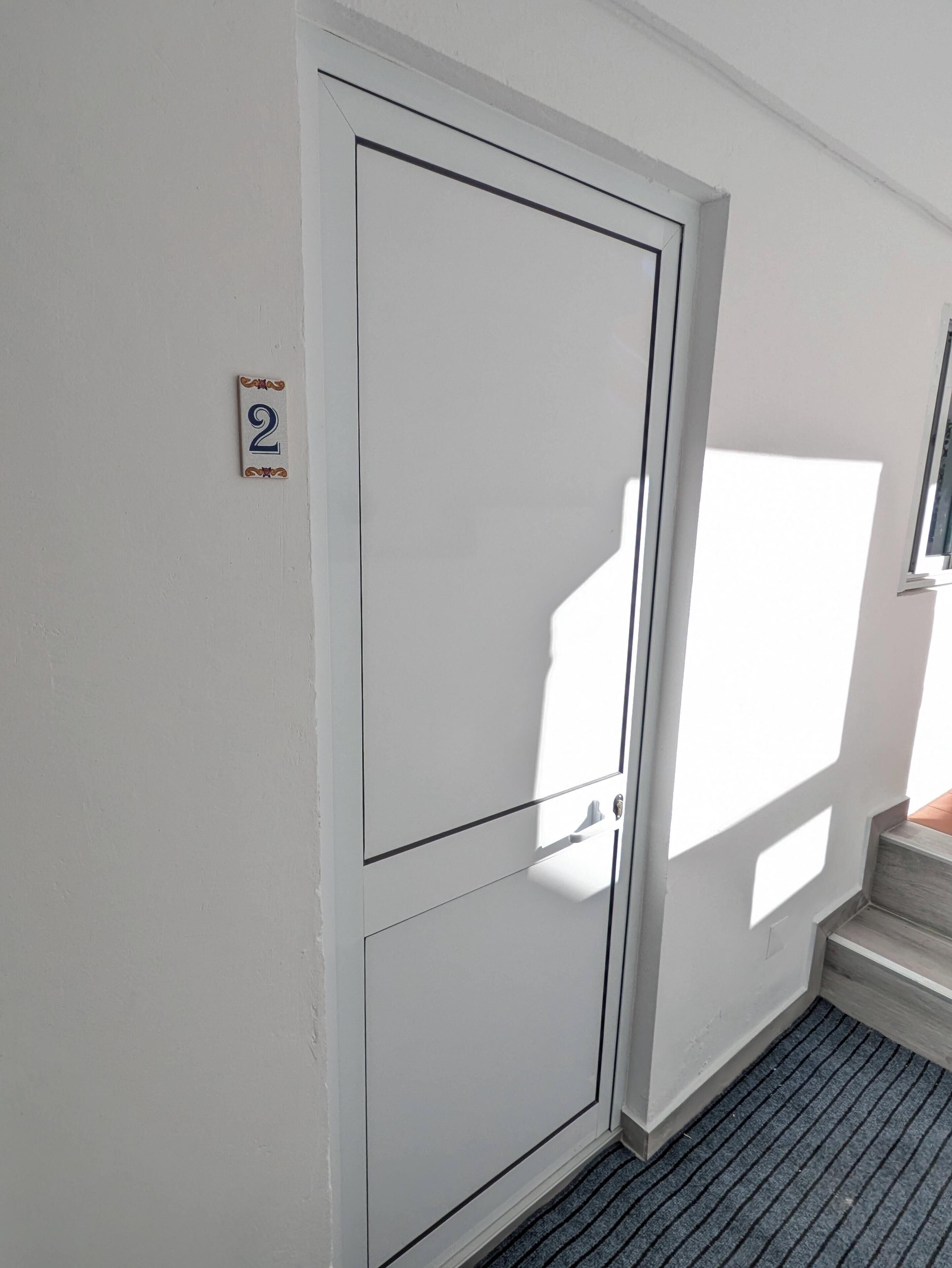 A white door with a number '2' sign, a window casting a shadow, and a small set of steps leading to a door.