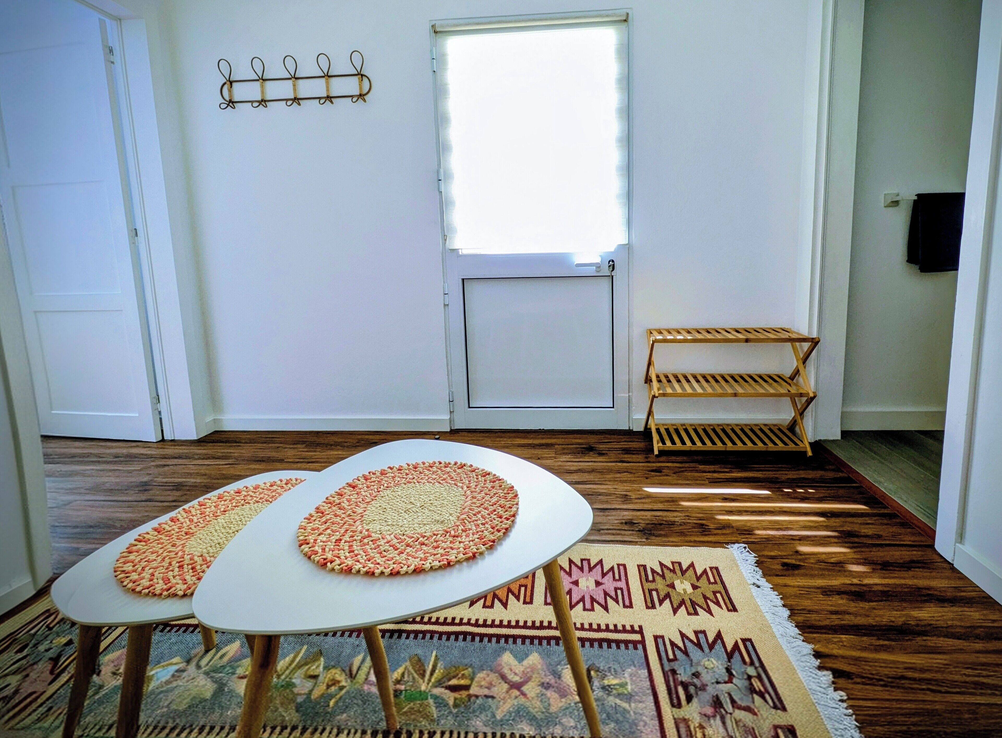 Una habitación con una puerta blanca, una ventana con persianas, un pequeño estante de madera, dos mesas redondas con individuales tejidos y una alfombra estampada en el suelo.