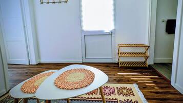 Una habitación con una puerta blanca, una ventana con persianas, un pequeño estante de madera, dos mesas redondas con individuales tejidos y una alfombra estampada en el suelo.