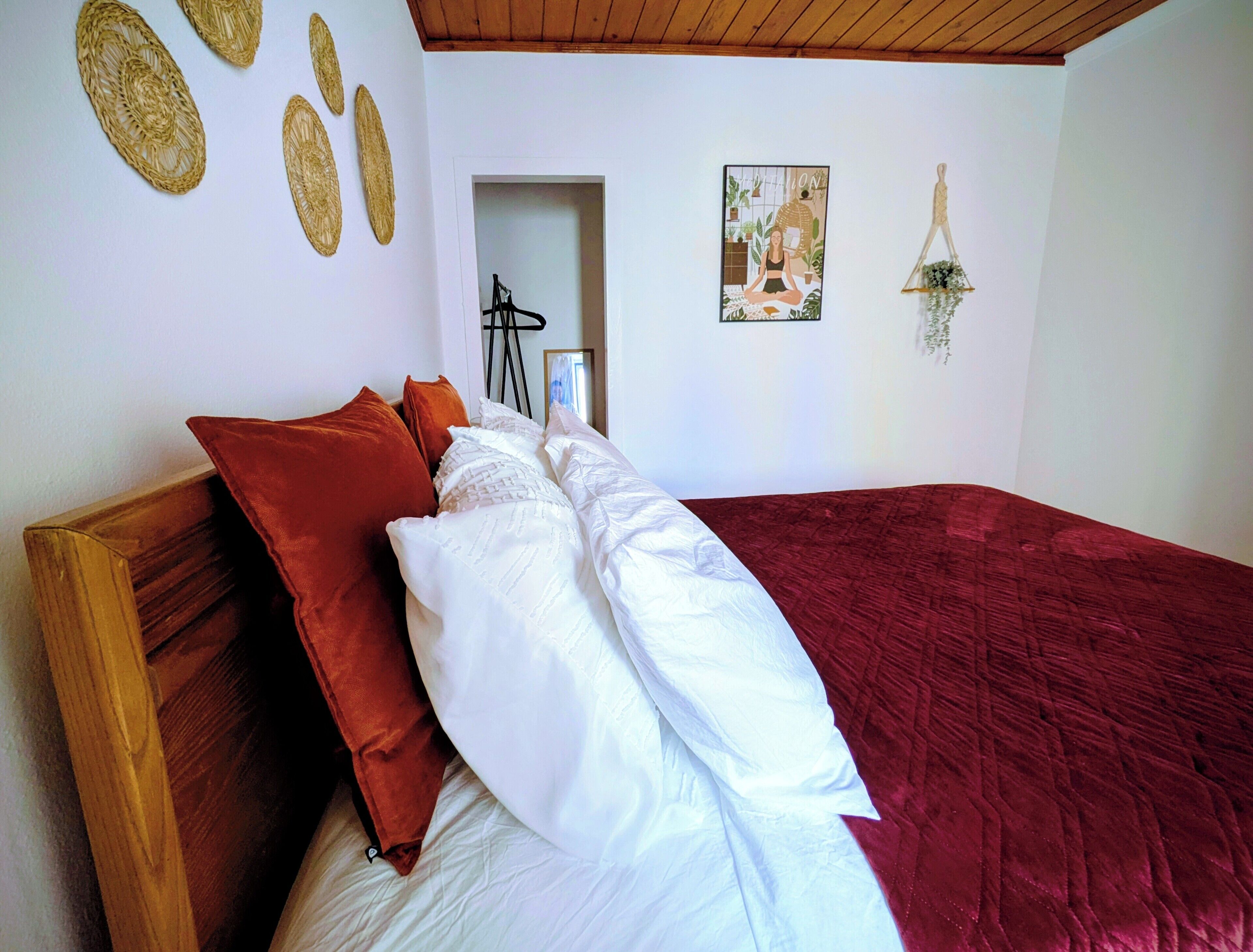 Un dormitorio con estructura de cama de madera, ropa de cama blanca y roja, colcha roja y adornos colgantes en la pared.