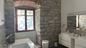 A bathroom with a stone wall, a window with blinds, a bathtub, and a sink with a mirror.