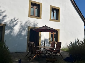 Un patio con una mesa, sillas y un paraguas pequeño, junto a un edificio blanco con dos ventanas.