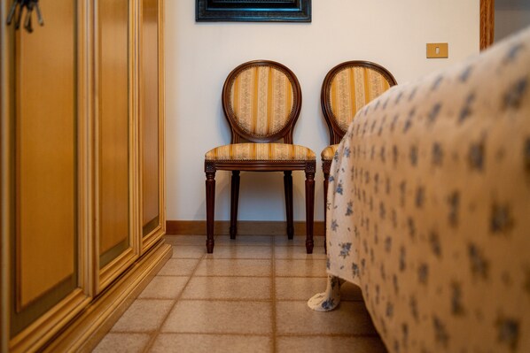 Habitación de hotel con una cama, dos sillas de madera tapizadas con tela a rayas, piso de baldosas e interruptor de luz en la pared.
