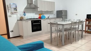 Eine kleine Küche mit weißen Schränken, einem Gasherd und einem eingebauten Backofen. Ein Esstisch mit vier Stühlen und ein kleiner Fernseher auf einem Holzgestell.