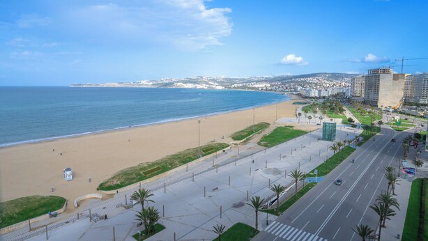 Un paysage urbain côtier avec une plage, une route bordée de palmiers et des bâtiments en arrière-plan.