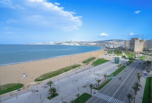 A coastal cityscape with a beach, a road with palm trees, and buildings in the background. - 2-Bedroom Apartment with Stunning Sea View in Tangier (Tangier)