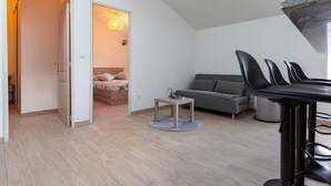 A modern, compact living space with a loft-style ceiling, a grey sofa, a small table, and a bar area with stools.