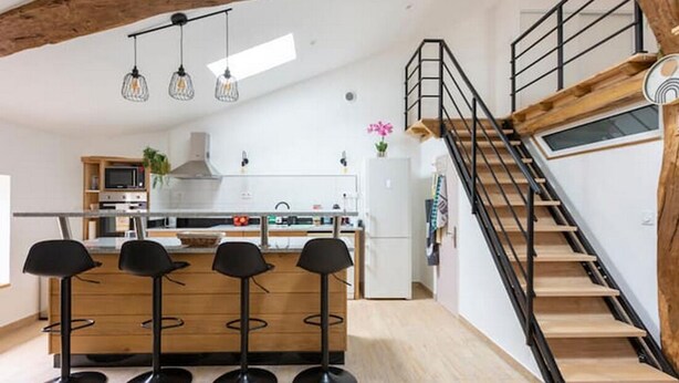 A modern kitchen with a central island, bar stools, a staircase, and a wooden ceiling.
