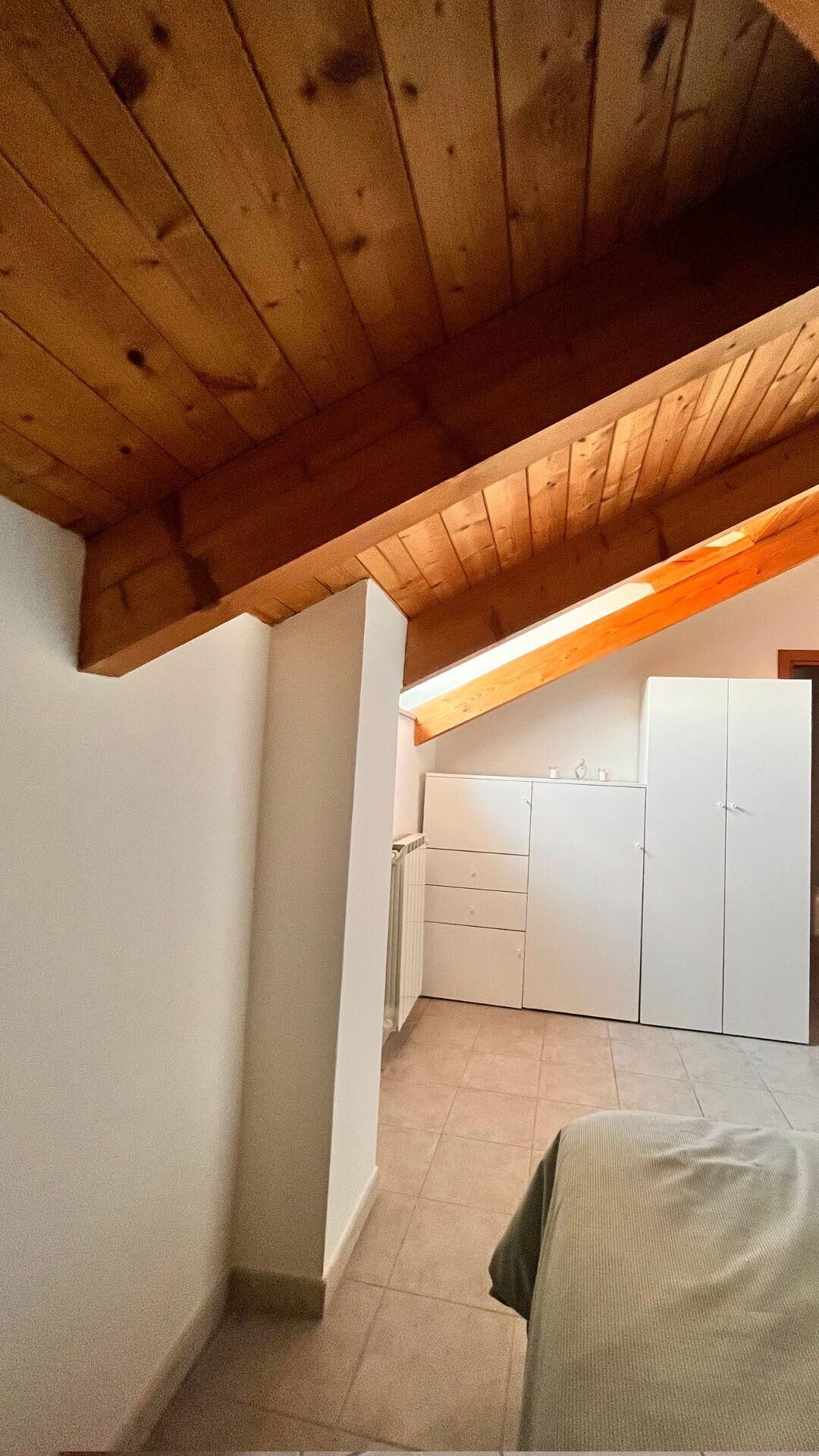 A room with a sloped ceiling, white walls, and a tiled floor. There is a white wardrobe and a small kitchenette area with cabinets.