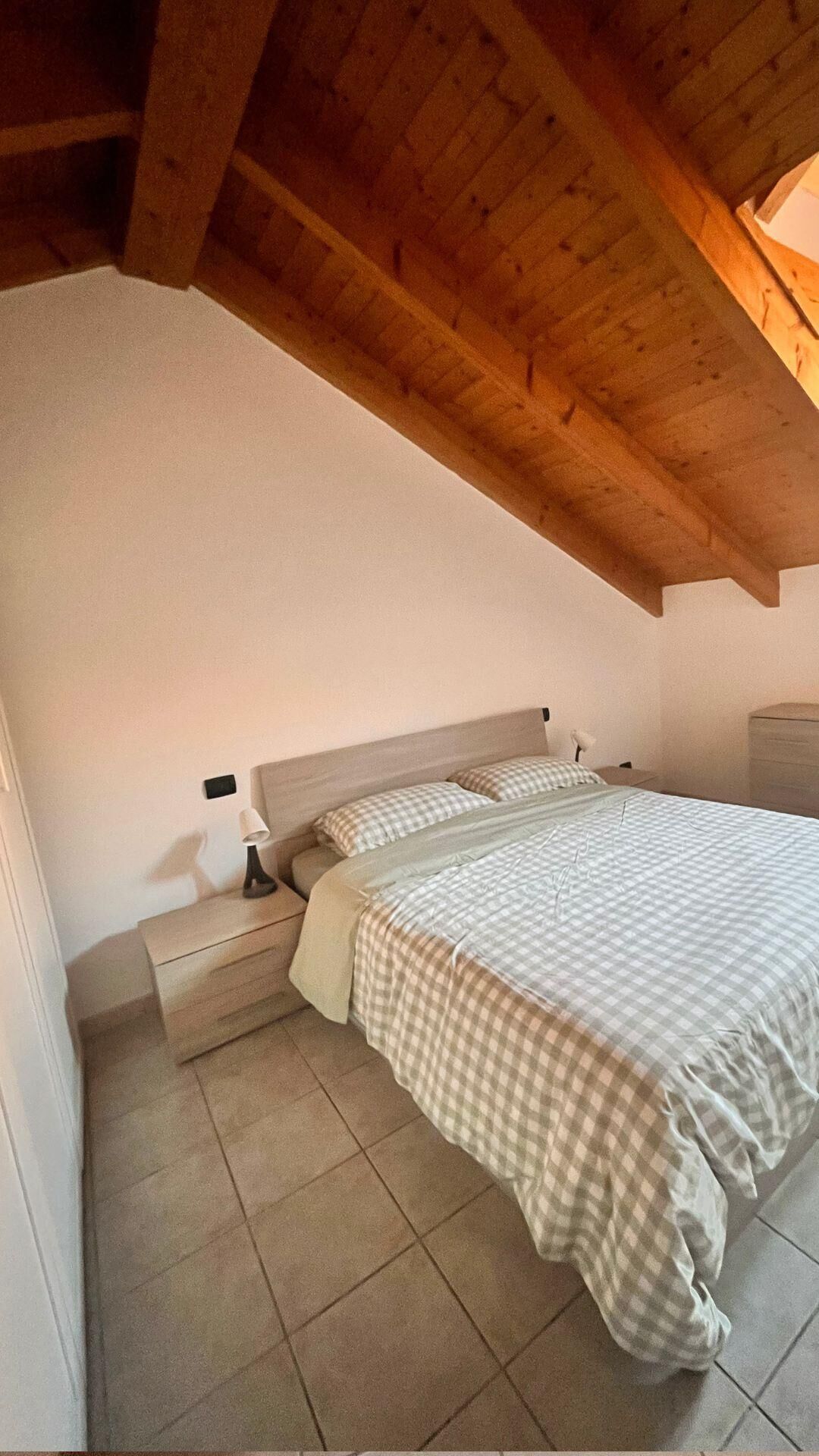 Une chambre avec un plafond en bois, un lit avec une couette à carreaux, une table de chevet et une lampe.