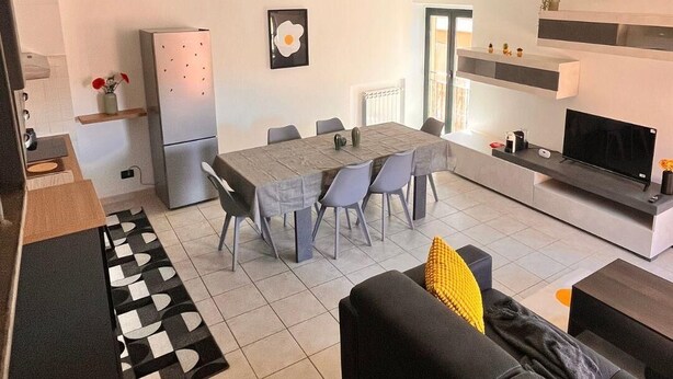 A modern living room with a sofa, a dining table with chairs, a kitchen area with a refrigerator, and a TV. - Hostpitaly   La Quiete in Centro (CHIVASSO)