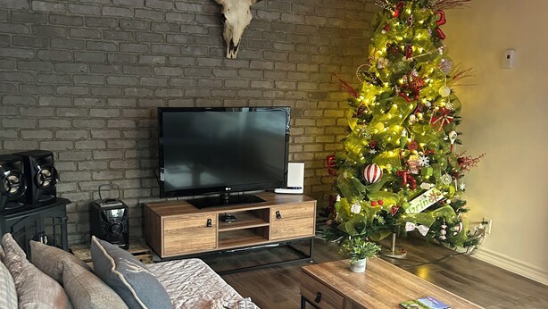A living room with a brick wall, a mounted TV, a Christmas tree, a sofa, and a coffee table. - Le petit chalet Chertsey (Chertsey)