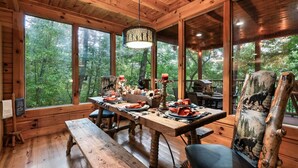 Un chalet rustique en bois, doté d’une table à manger pour quatre personnes, de grandes fenêtres donnant sur la forêt et d’un luminaire suspendu.