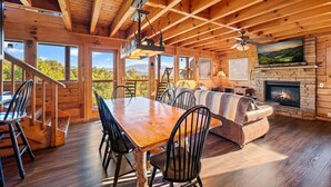 A wooden cabin interior with a dining area, sofa, fireplace, and large windows offering scenic views. - 4BR w/Mountain Views! Hot Tub*Game Room*Comm Pool (Sevierville)