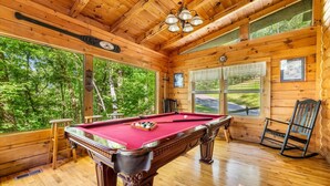 Une table de billard dans un chalet en bois doté de grandes fenêtres et offrant une vue sur la forêt.