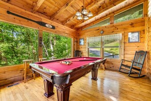 Une table de billard dans un chalet en bois doté de grandes fenêtres et offrant une vue sur la forêt.