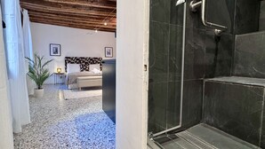 A hotel room with a bed, a bedside table with a lamp, a potted plant, and a shower area with dark tiles. - Mozart Bis (5 min to Rialto's Bridge) (Venecia)
