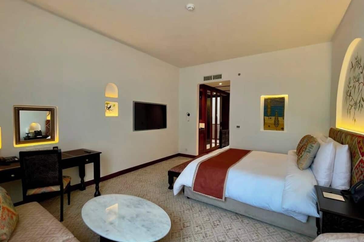 A hotel room with a bed, a desk, a chair, a small table, and a TV.