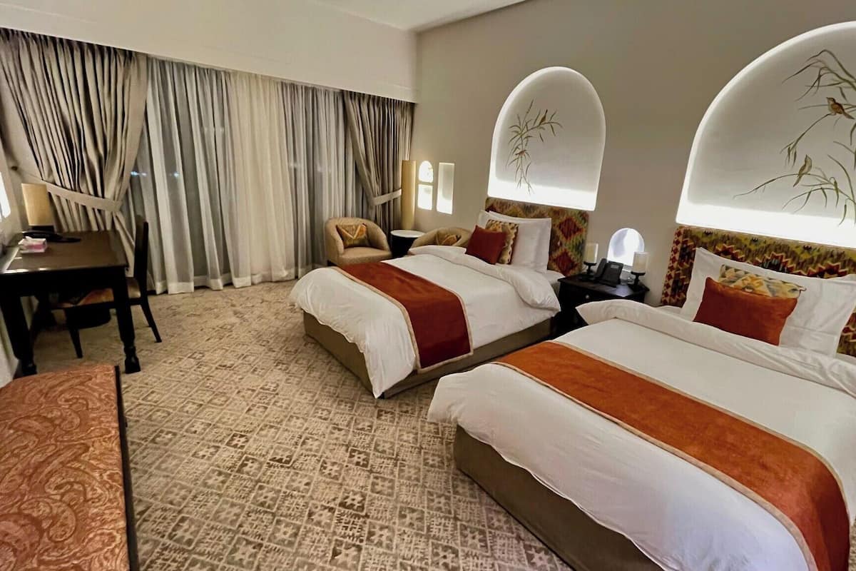 A hotel room with two beds, a desk, a chair, and a large window with curtains.