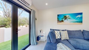 A modern living room with a large window, a grey sofa, a small white side table, and a beach-themed painting on the wall. - Palm Beach Hideaway #8 - New Construction 2Bedrooms 2bathrooms Pool/Rooftop (Noord)