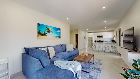 A modern living room with a blue sofa, a coffee table, and a painting of a beach scene. - Palm Beach Hideaway #8: Poolfront Corner 2BR with Wraparound Patio (Noord)
