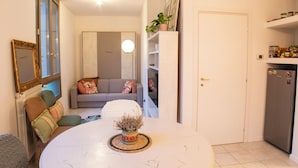 A modern living room with a grey sofa, a round coffee table with a decorative plant, and a white door leading to a kitchen with a refrigerator. - Conti & Moroni House  (Calenzano)