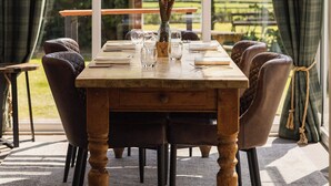 A dining area with a wooden table, six chairs, and a view of a garden through large windows. - The Haughmond (Upton Magna)