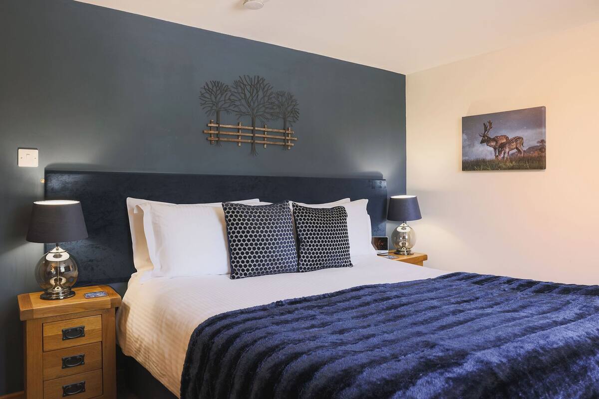 Ein Schlafzimmer mit einem Bett, zwei Nachttischen mit Lampen, einer Kommode mit Schubladen, einem dunkelblauen Kopfteil, einem Wandbild mit Hirschen und einer Wanddekoration mit einem Baum.