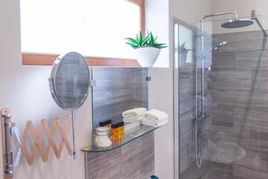 A modern bathroom with a glass shower enclosure, a wooden towel rack, a glass shelf with towels, a round mirror, and a potted plant.