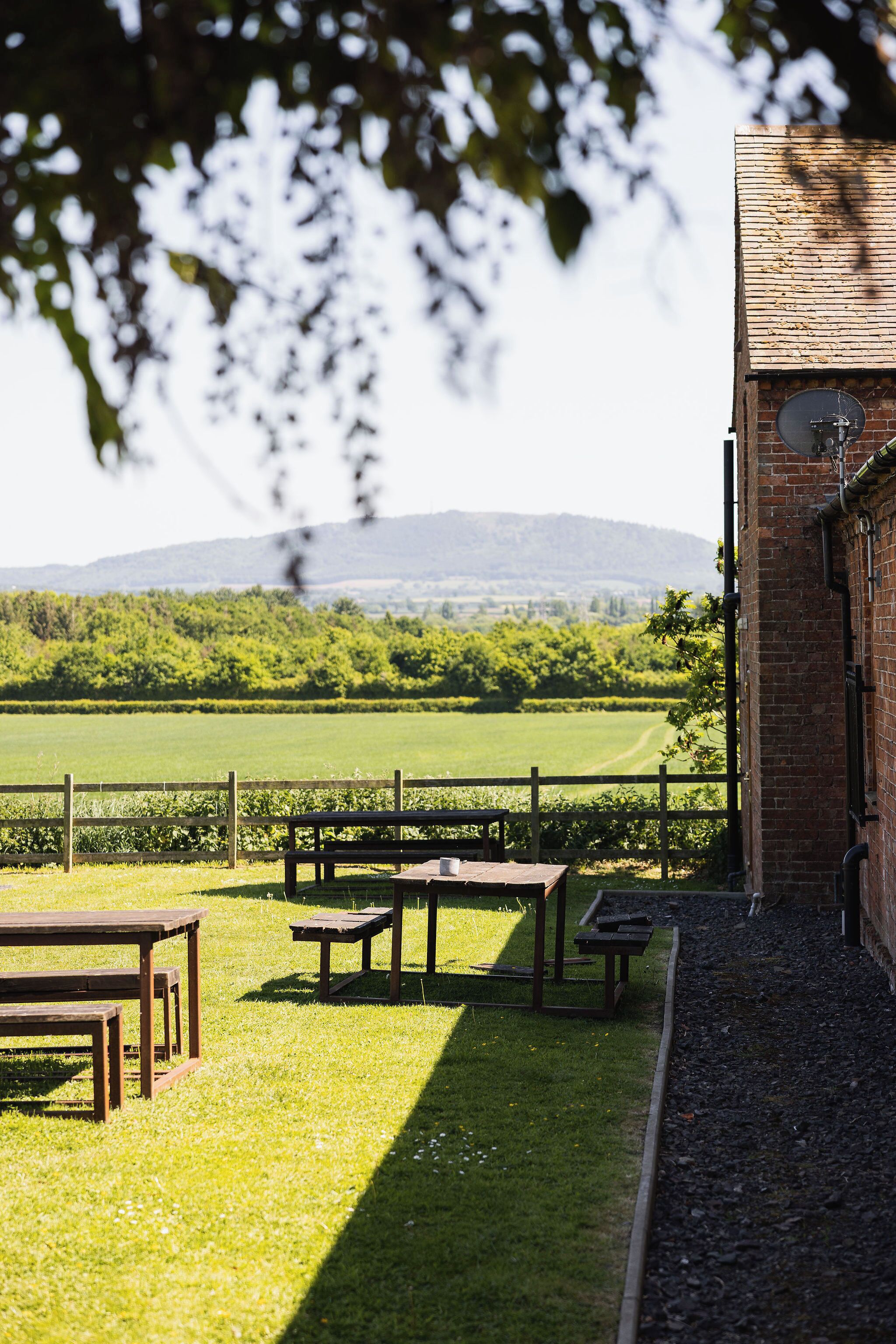 An outdoor seating area with picnic tables and a view of a green field and mountains in the distance.