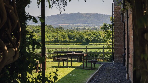 An outdoor area with a pathway leading to a seating area, surrounded by greenery and a view of distant hills. - The Haughmond (Upton Magna)