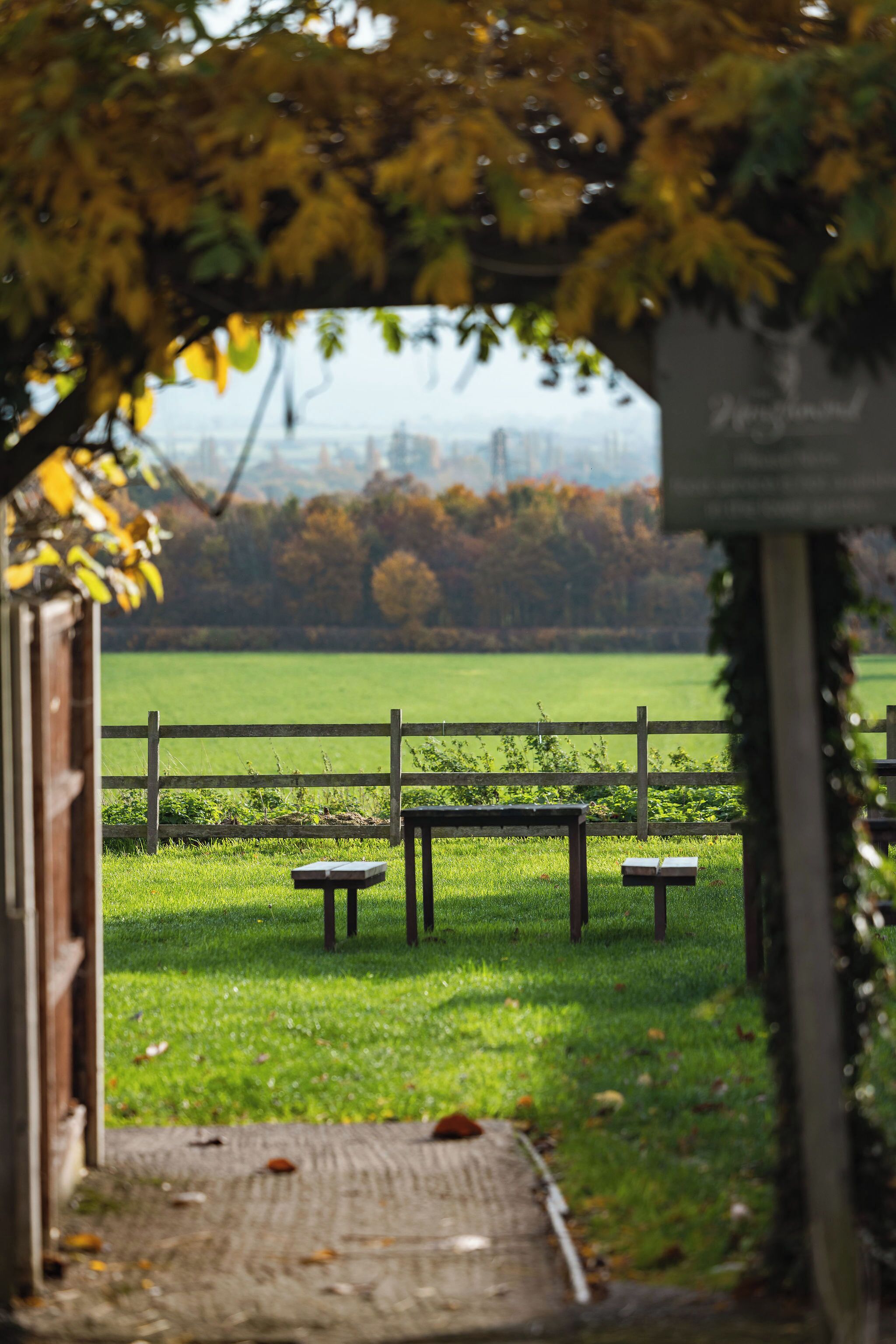 An outdoor seating area with tables and benches under a wooden archway, leading to a grassy field with a fence in the background.