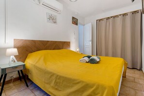 A hotel room with a bed, a bedside table with a lamp, a wall-mounted air conditioner, and a door with a curtain. - Gîte Les Garrigues – Modern Comfort Near Les Baux-de-Provence, Sleeps 3 (Mouriès)