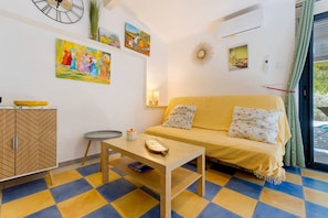 A living room with a sofa, a table, and a cabinet. There are paintings on the wall and an air conditioning unit. - Gîte Les Garrigues – Modern Comfort Near Les Baux-de-Provence, Sleeps 3 (Mouriès)