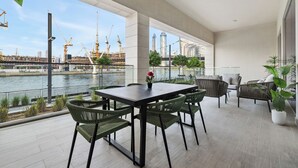 A modern outdoor dining area with a view of a river and construction cranes in the background. - 2BR M Huge Terrace Direct Canal Access (Dubai)