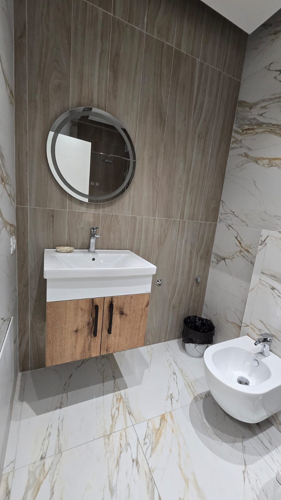 Un baño con tocador de madera, un espejo redondo, un lavamanos y un bidé.