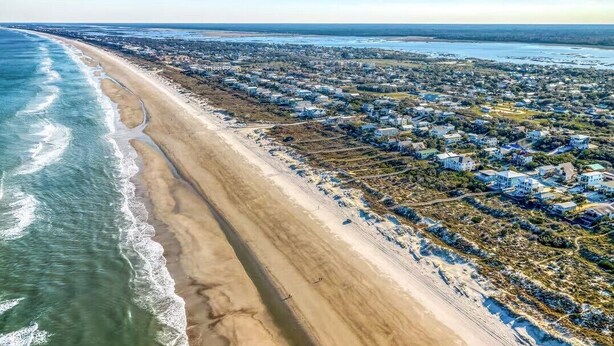 Vista aérea de uma área residencial costeira com uma extensa praia de areia, casas e uma visão clara do oceano.