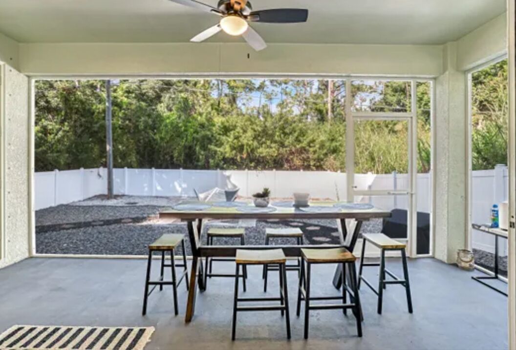 A sunlit room with a table and chairs, a ceiling fan, and a view of a backyard with trees and a white fence.