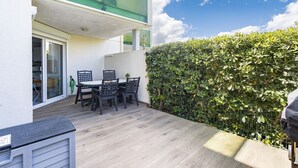 A patio area with a table and chairs, a glass door leading to an interior space, and a hedge along the side. - Seaside Apartment in Saint-Cyprien-Plage (66750), Lagune Area (Saint-Cyprien Plage)