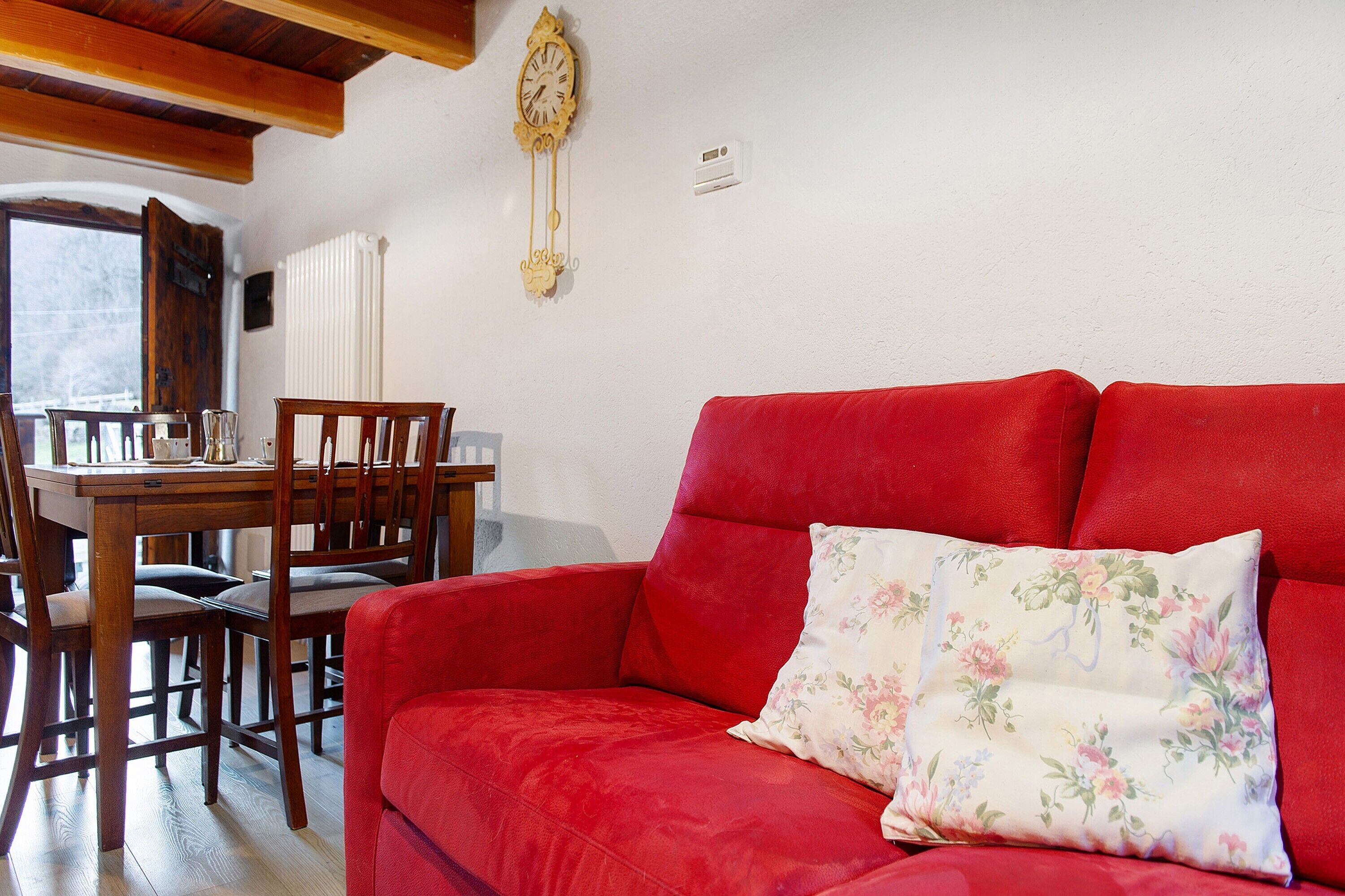 A red sofa with a floral pillow, a wooden dining table with chairs, and a wall-mounted clock.