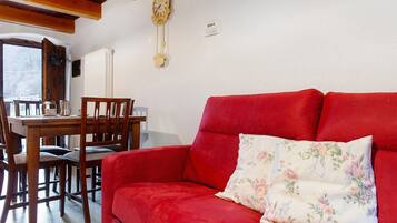 A red sofa with a floral pillow, a wooden dining table with chairs, and a wall-mounted clock.